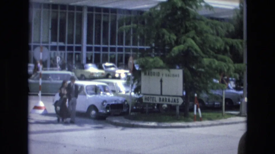 1972 Spain street scene showing a couple with luggage near a white compact car, Hotel Barajas signage, parked cars, trees, and a glass-fronted building.