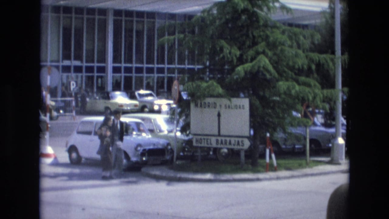 In Spain in 1972, a couple with bags crosses a busy street near a sign for Madrid, Salidas, and Hotel Barajas, with a white Mini and other cars nearby.