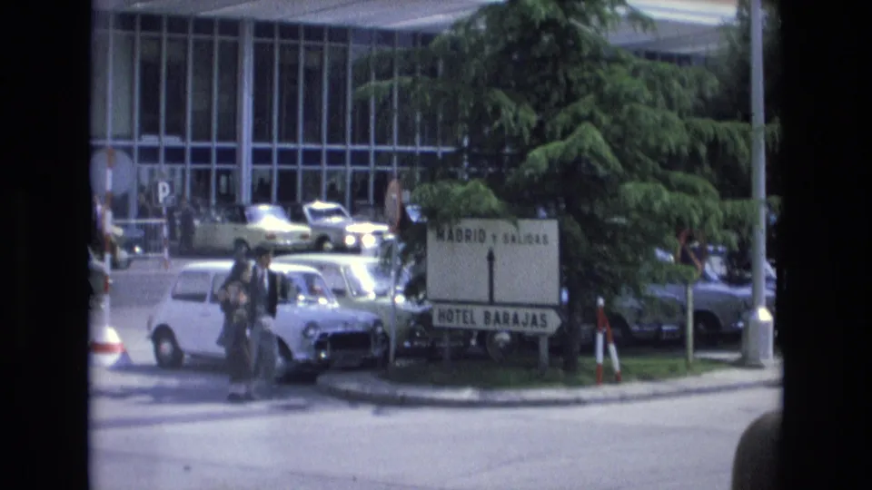 In Spain in 1972, a couple with bags crosses a busy street near a sign for Madrid, Salidas, and Hotel Barajas, with a white Mini and other cars nearby.