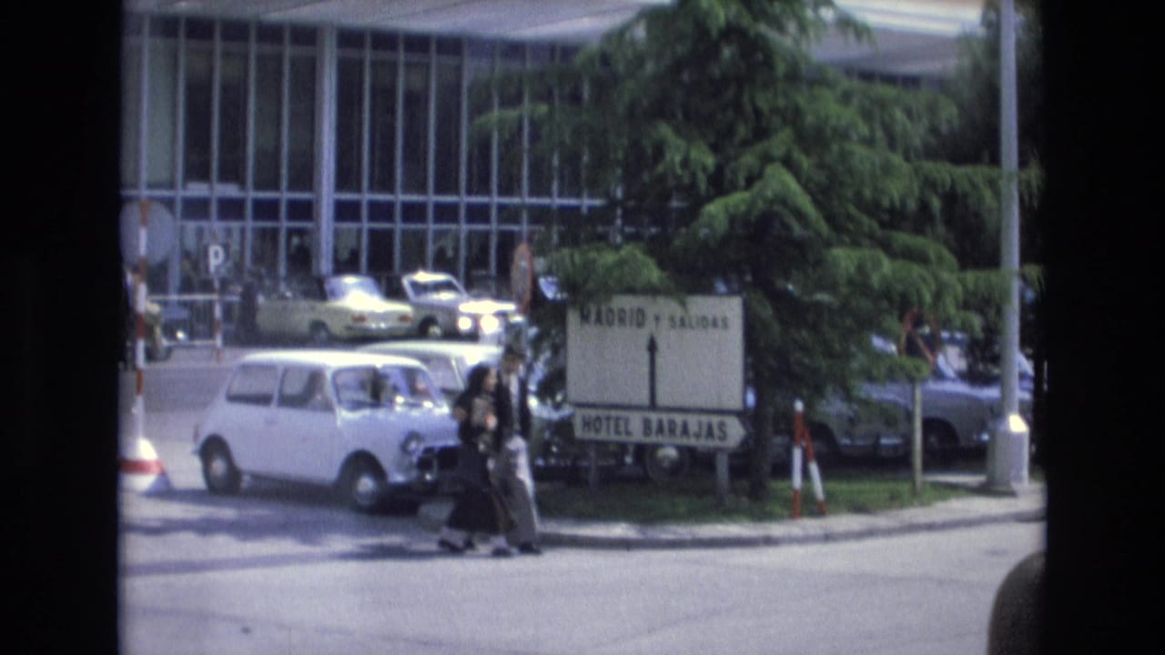 Two pedestrians walk past a road sign as cars move near a modern glass-fronted building.