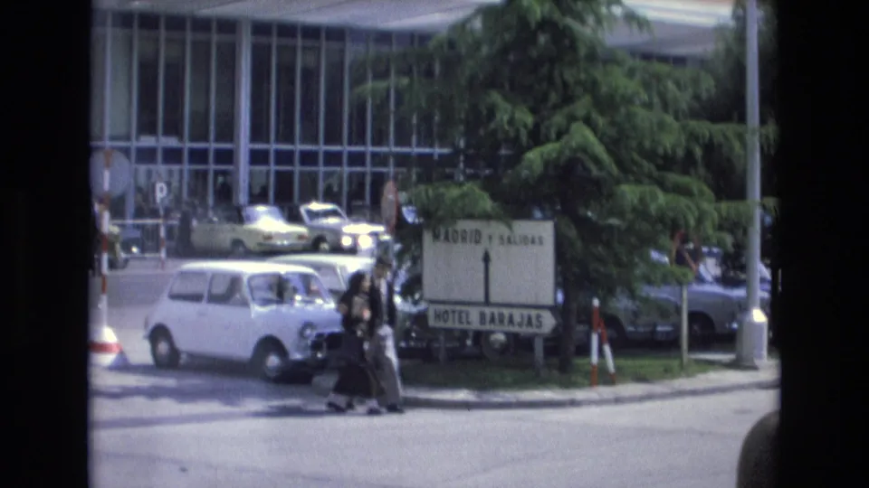 In the 1972 Spain street scene, two pedestrians pass the Hotel Barajas sign while traffic and parked cars sit in front of a large glass-windowed building.
