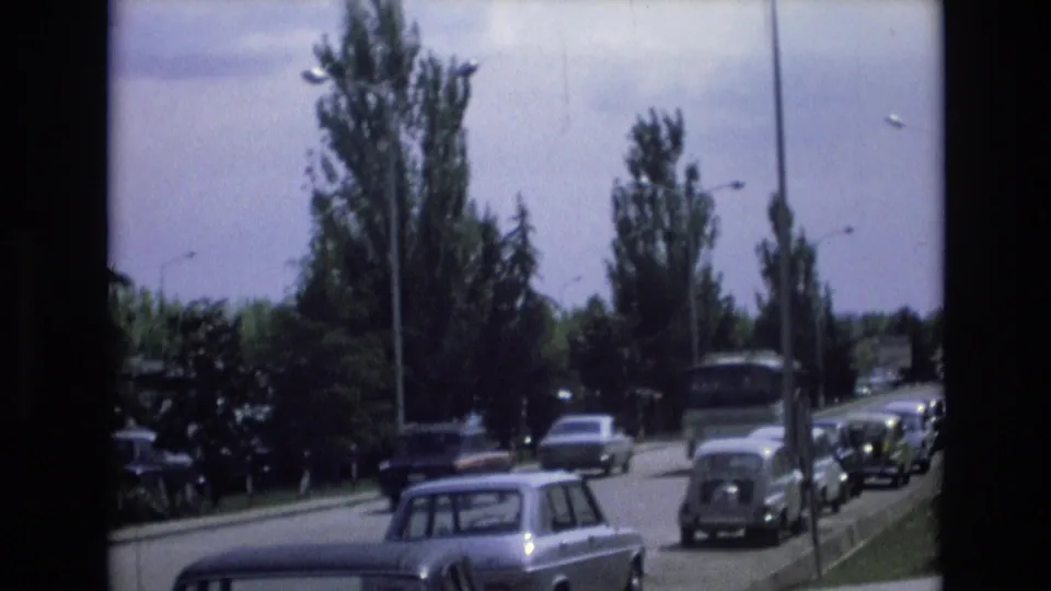 The 1972 Spain footage shifts to a handheld view down a roadside lined with parked cars, tall trees, lampposts, and passing traffic.
