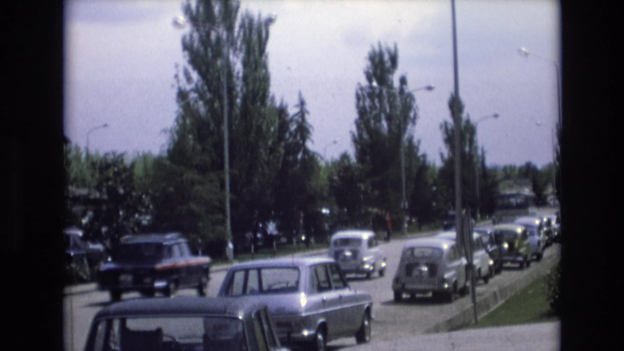 A dark car passes on a suburban road while other cars line the curb.