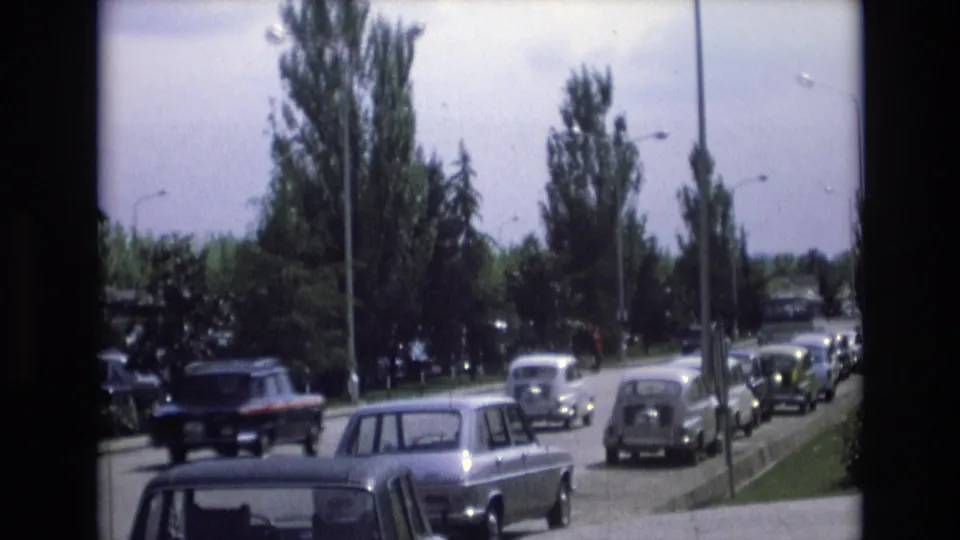 1972 Spain 8mm frame with a dark car moving through a line of parked and approaching vehicles on a suburban tree-lined street.