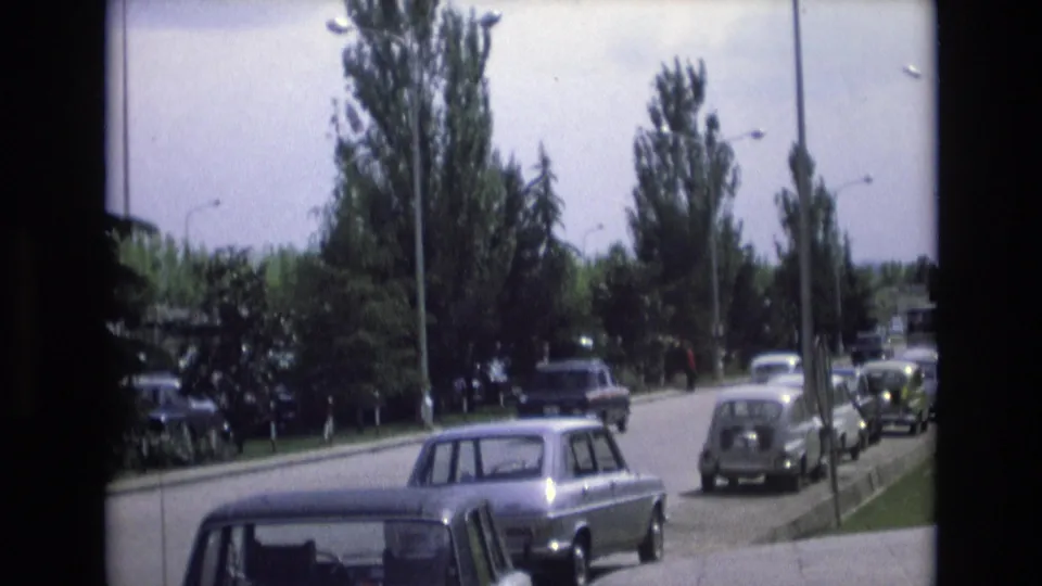 1972 Spain 8mm frame showing a suburban street with classic European cars, tall trees, lampposts, and a curbside parking lane.