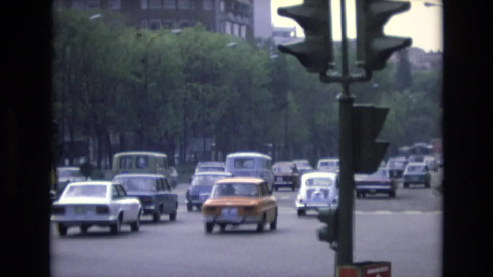1972 Spain street traffic seen through an 8mm home-movie frame, with compact cars, a foreground traffic signal, trees, and urban buildings.