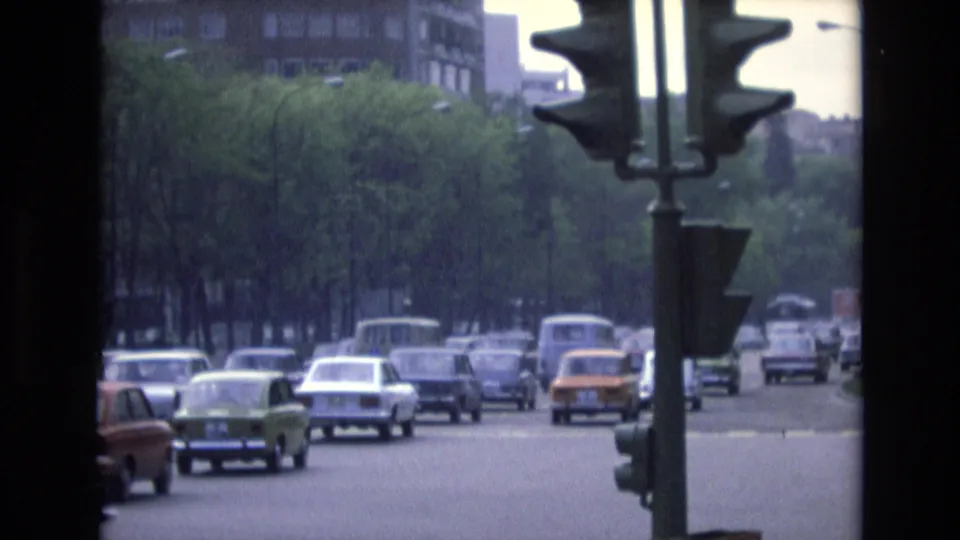 1972 Spain 8mm view of commuter cars passing through an urban roadway, with a large traffic signal, green trees, and distant vehicles.