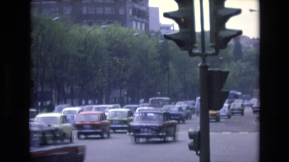1972 Spain home-movie frame showing heavier urban traffic with compact sedans, a traffic light, tree cover, and mid-rise buildings in the background.