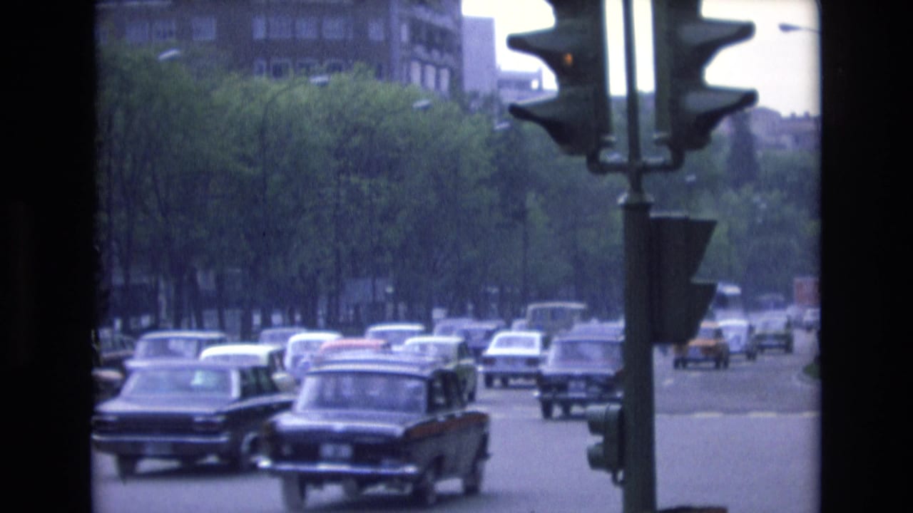 1972 Spain 8mm frame showing period cars moving through a tree-lined city street, with a traffic signal and distant buildings visible through dark film borders.