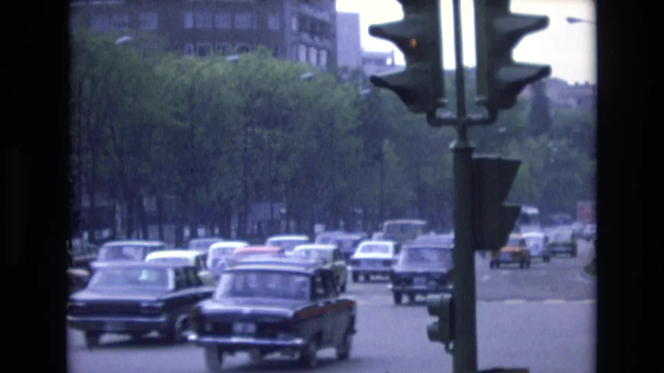 1972 Spain 8mm frame showing period cars moving through a tree-lined city street, with a traffic signal and distant buildings visible through dark film borders.