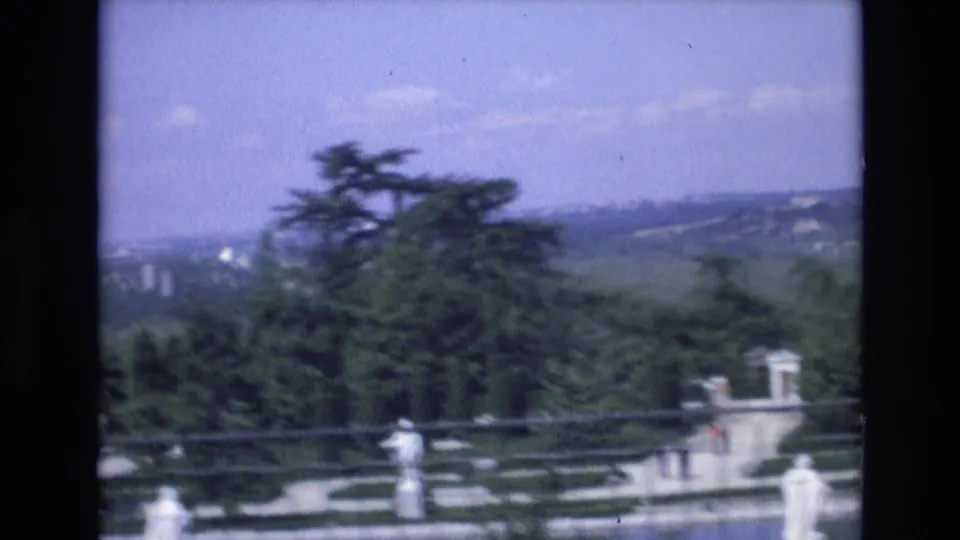 1972 Spain 8mm frame from a scenic overlook, showing formal garden paths, white statues, evergreen trees, and a hazy city landscape in the distance.