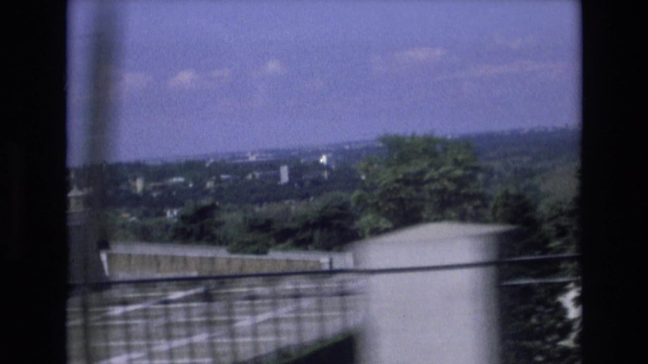 1972 Spain 8mm travel frame showing a distant rural landscape, roadside barrier, trees, utility line, and cloudy sky from a moving vehicle.