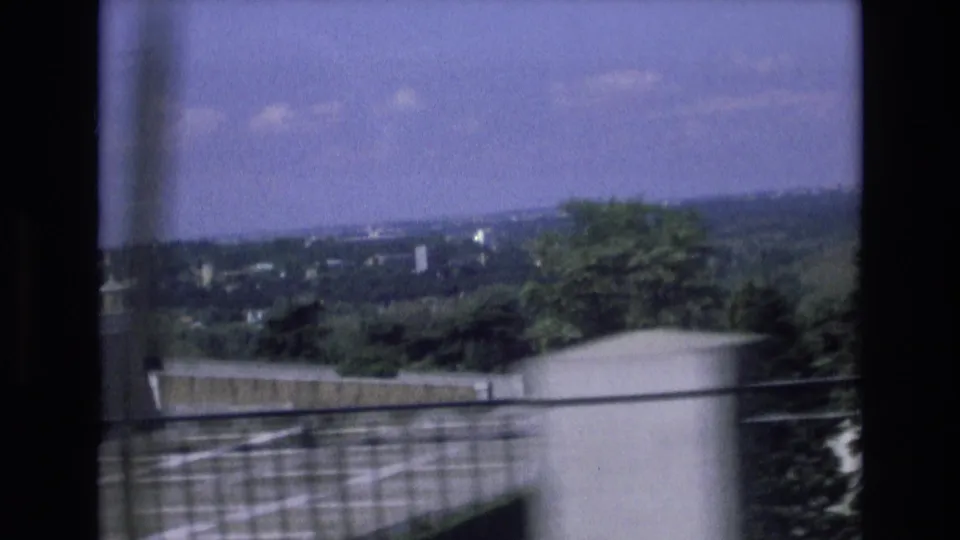 1972 Spain 8mm travel frame showing a distant rural landscape, roadside barrier, trees, utility line, and cloudy sky from a moving vehicle.
