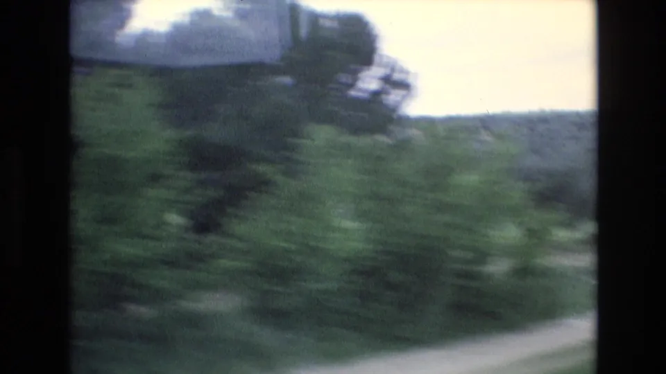 1972 Spain 8mm frame from a moving car showing a blurred green slope, trees, distant pale object, and bright sky at the edge of the road view.