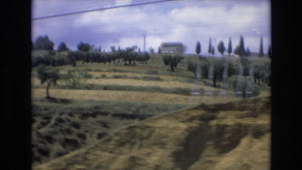 1972 Spain 8mm countryside view with open fields, rows of small trees, distant buildings, utility wires, and a partly cloudy sky filmed from a vehicle.