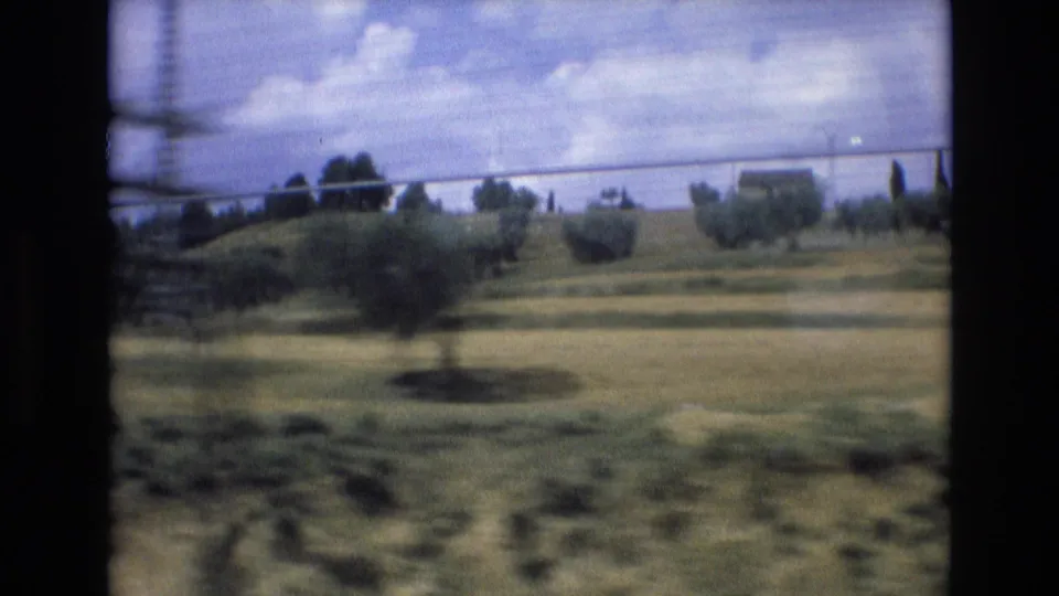 1972 Spain 8mm frame showing rural fields, scattered trees, utility wires, and distant low buildings viewed through motion blur from a moving vehicle.