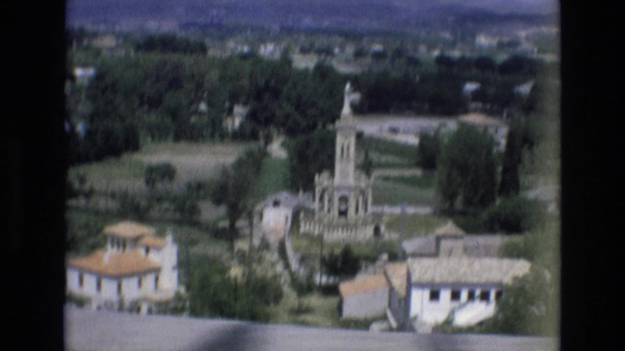 1972 Spain 8mm frame from an elevated viewpoint showing a town monument among trees, open fields, low buildings, and distant hills.