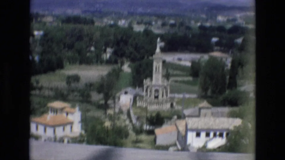 1972 Spain 8mm frame from an elevated viewpoint showing a town monument among trees, open fields, low buildings, and distant hills.
