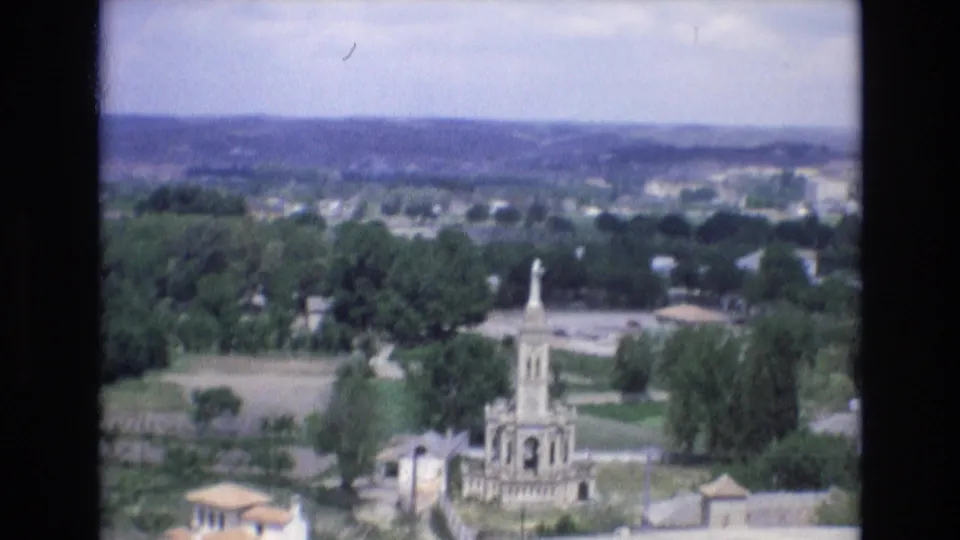 1972 Spain 8mm frame showing a prominent stone monument with a statue-like top, surrounded by trees, open land, and scattered town buildings.