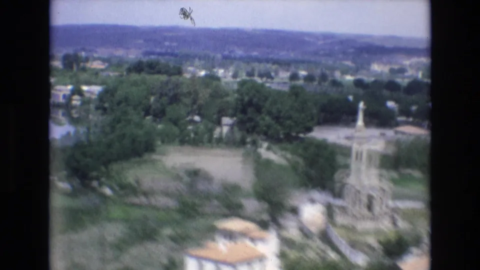 1972 Spain 8mm frame with motion softness as the camera passes over a stone monument, nearby rooftops, trees, fields, and the hilly background.