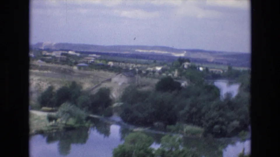 1972 Spain 8mm frame looking over a river bend, dense trees, open countryside, scattered buildings, and low hills under a bright sky.