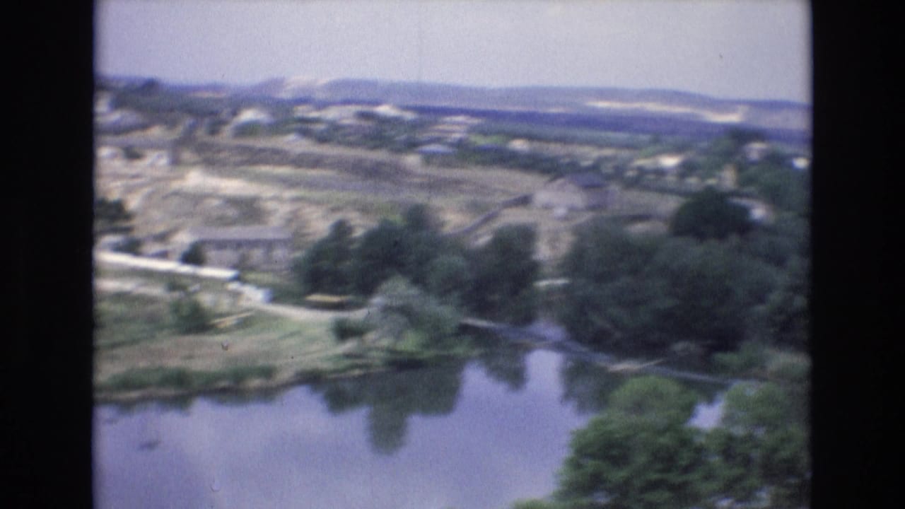 1972 Spain 8mm frame looking from an elevated route across a river or reservoir edge toward roads, trees, fields, and a small hillside town.