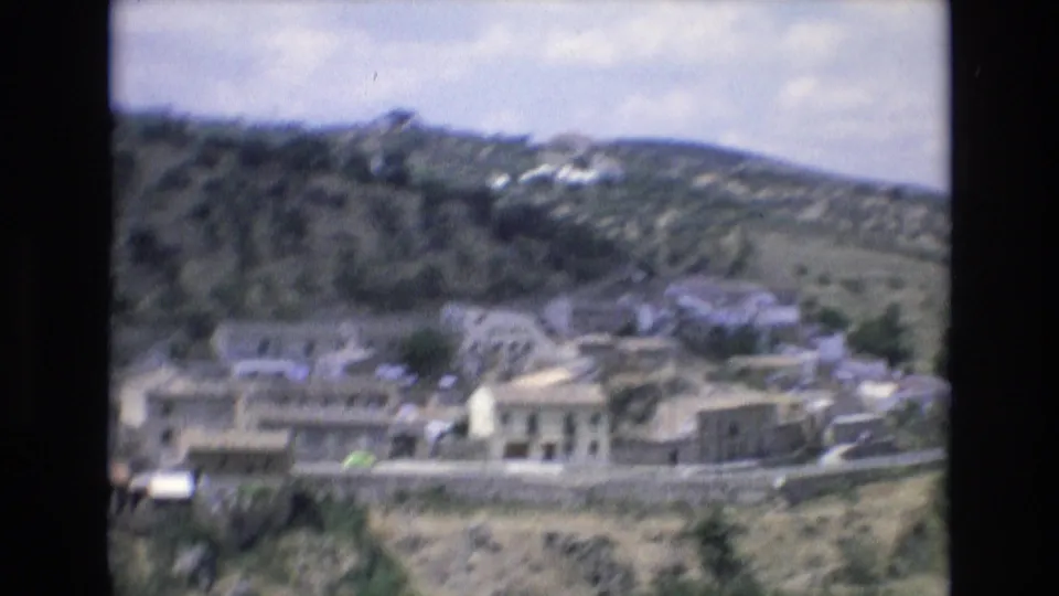1972 Spain 8mm frame showing clustered low buildings along a hillside road, with trees and higher ground rising behind them.