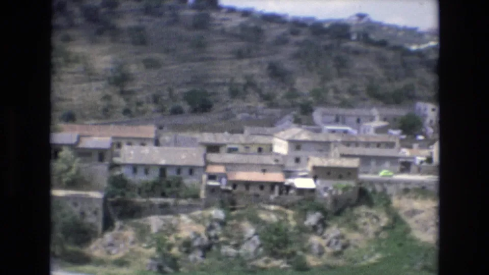 1972 Spain 8mm frame with low town buildings, a hillside road, rocky ground, trees, and terraced slopes seen from a moving elevated position.