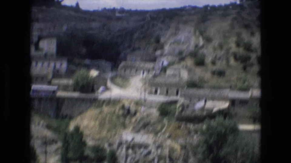 1972 Spain 8mm frame looking across an elevated roadway or bridge toward hillside buildings, trees, rocky slopes, and a small road below.
