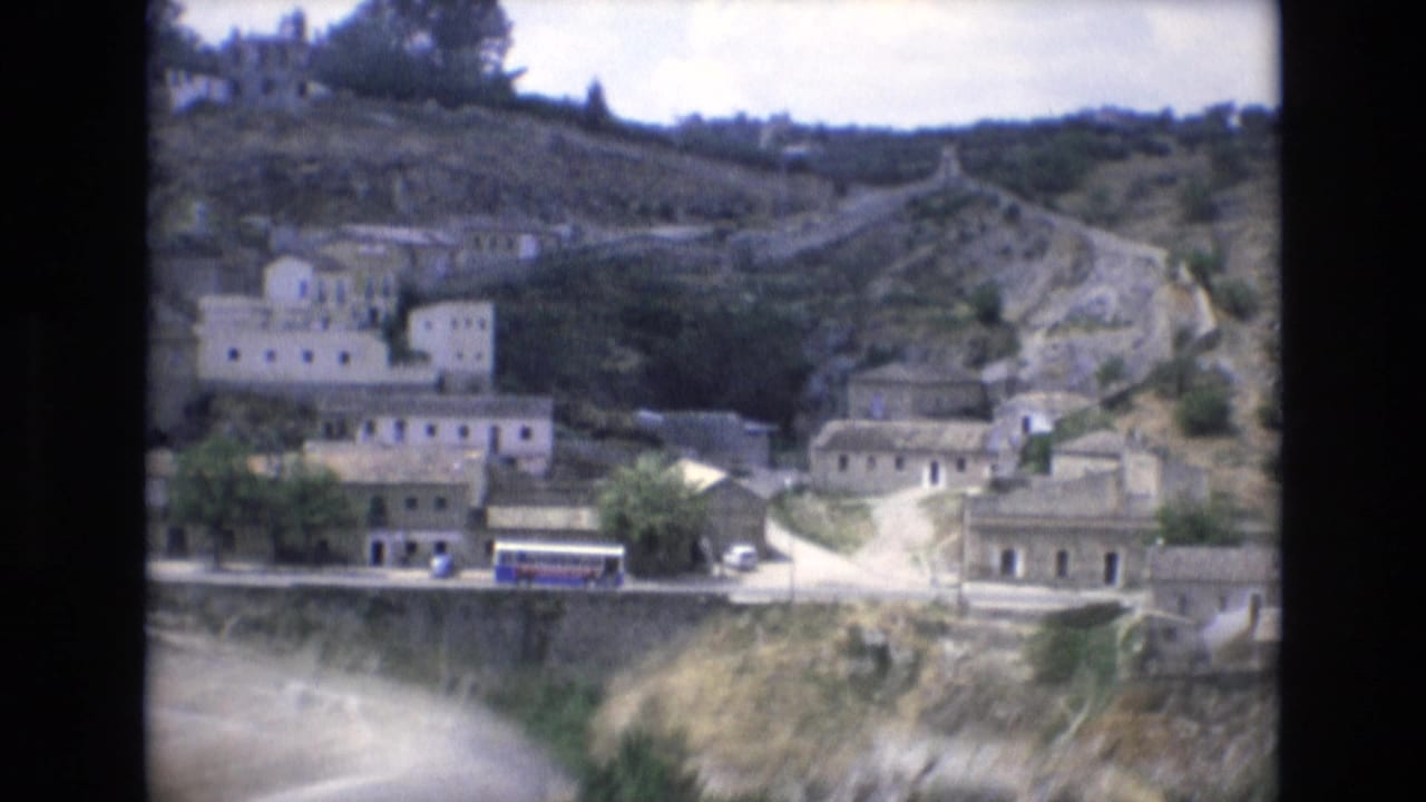 1972 Spain 8mm frame showing an elevated view over a rocky hillside settlement with white buildings, a winding road, and a blue bus along the lower edge.