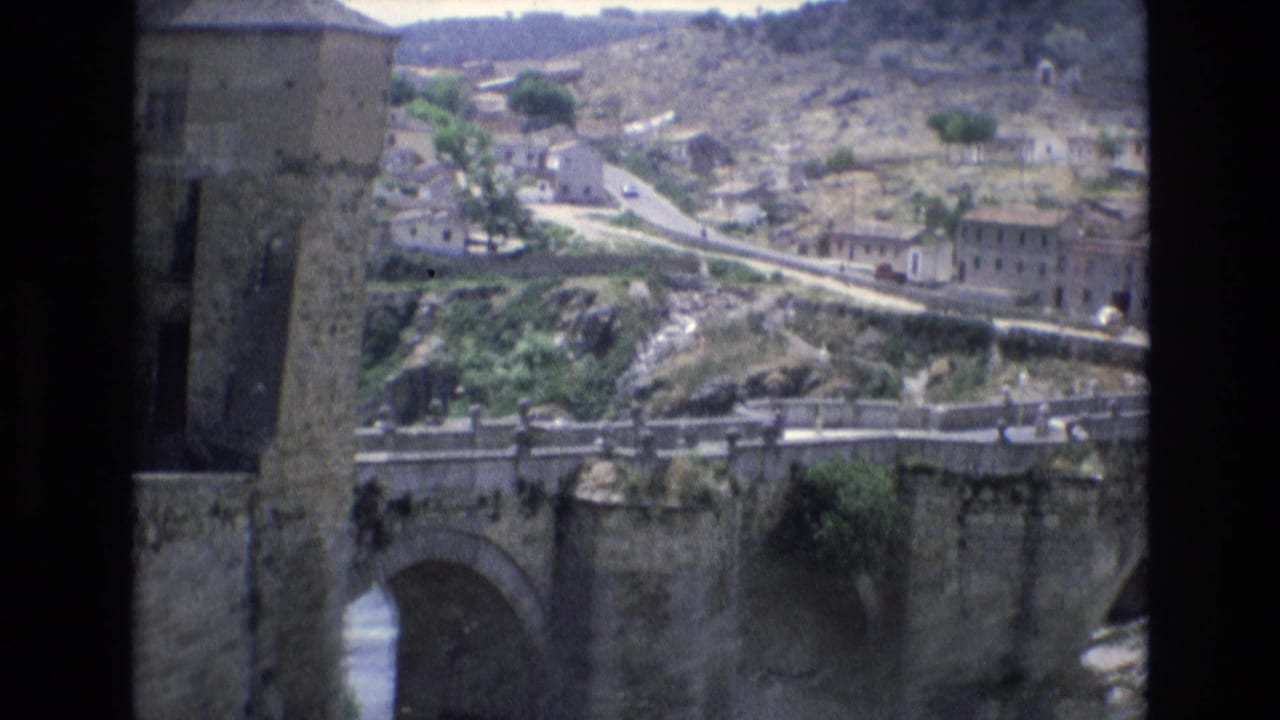 Stone arch bridge and hillside village seen from an elevated position beside a dark vertical structure.