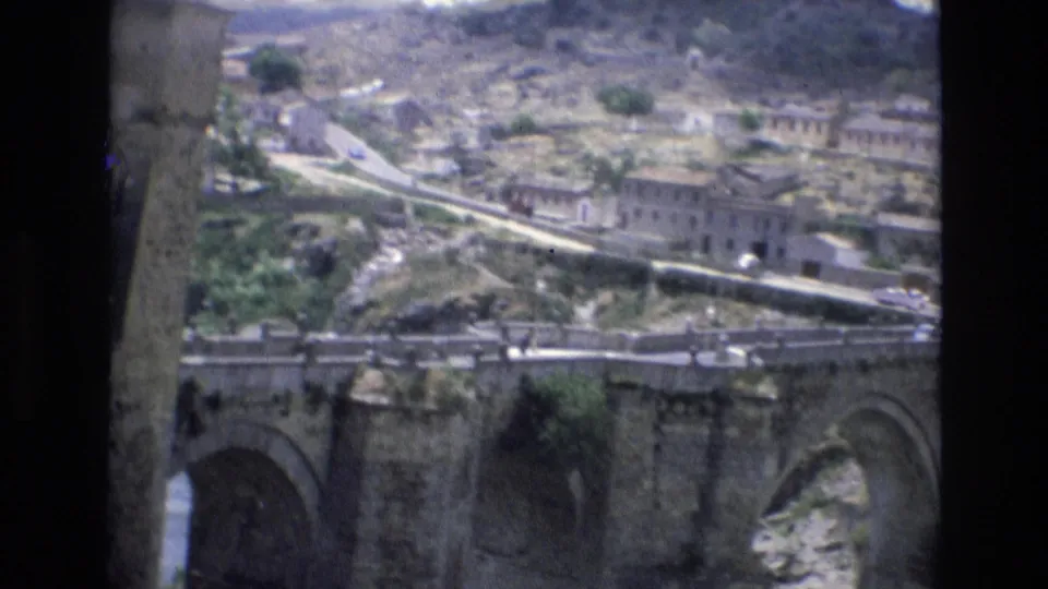 1972 Spain 8mm frame looking across an old stone bridge toward a road, rocky slope, and clustered hillside buildings in a historic town setting.
