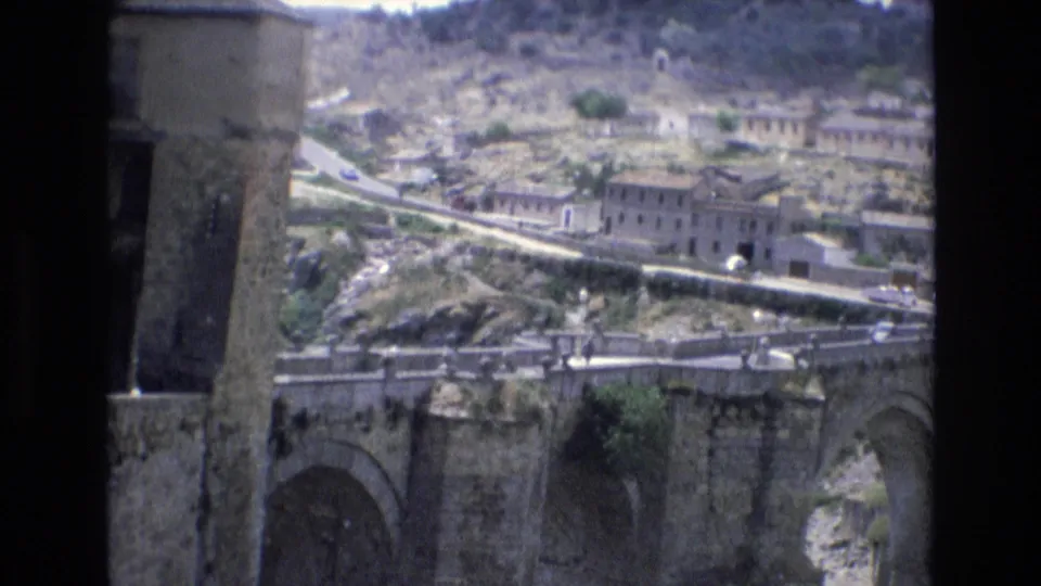 1972 Spain frame showing the stone bridge from an elevated visitor viewpoint, with hillside buildings, a road, and small vehicles visible beyond the masonry foreground.
