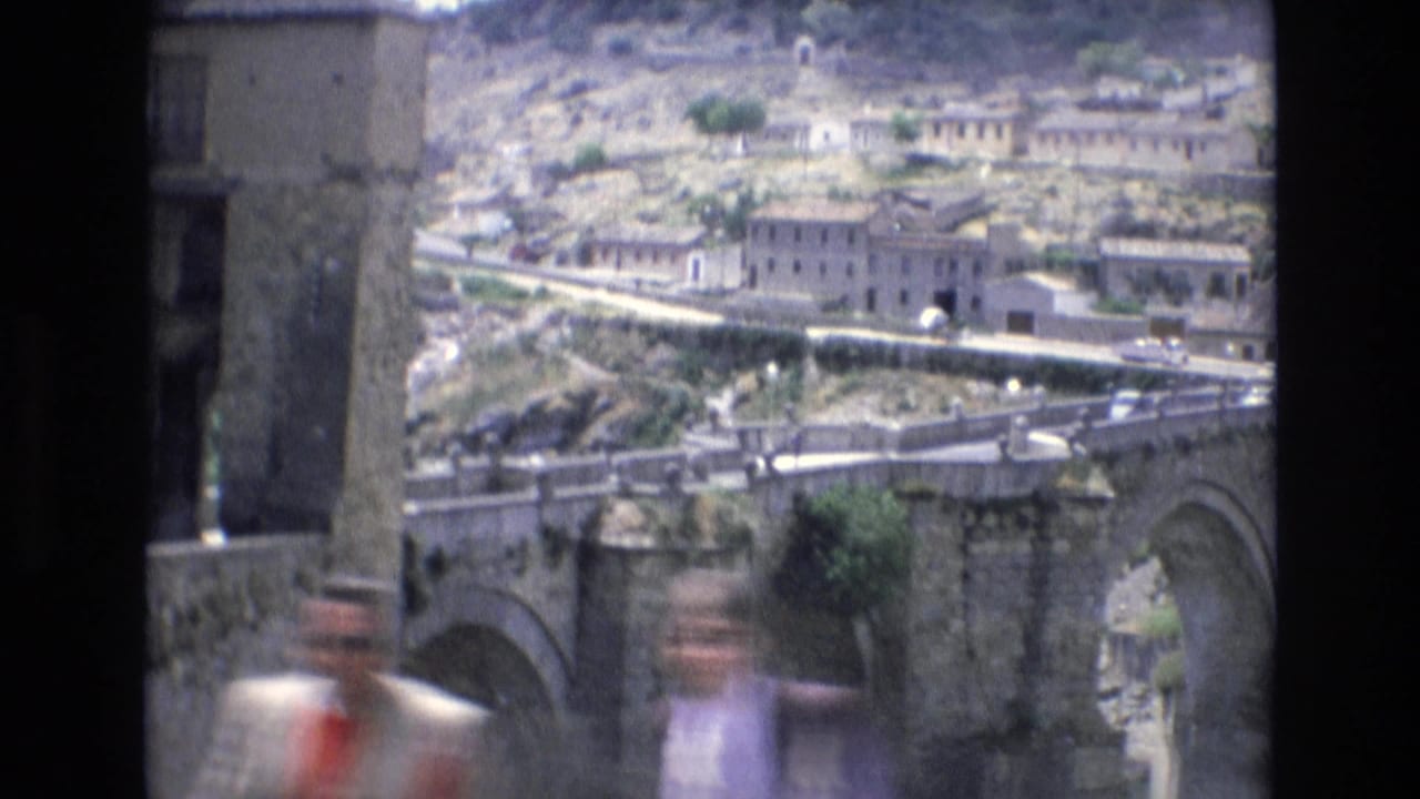 1972 Spain 8mm frame showing a historic stone bridge, hillside masonry buildings, a walled road, and blurred pedestrians passing close to the camera.