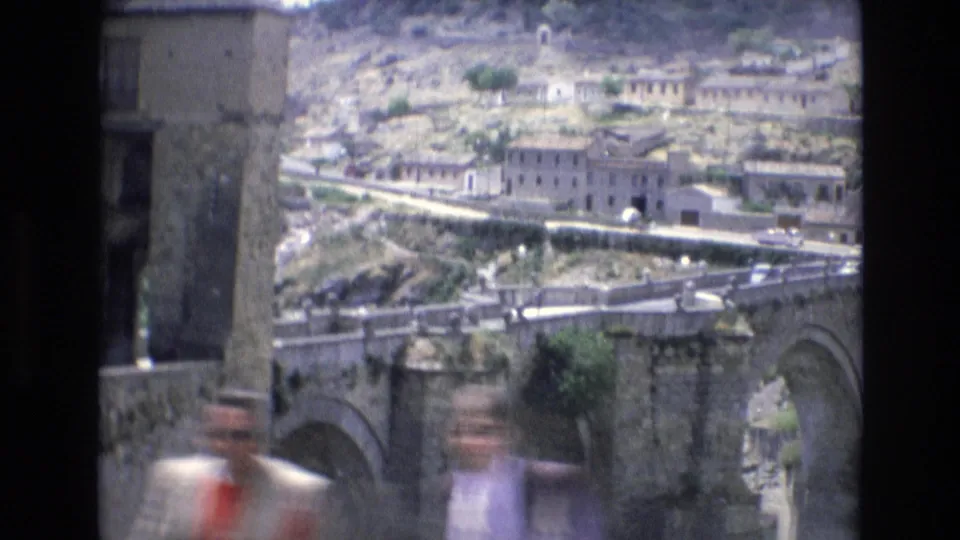1972 Spain 8mm frame showing a historic stone bridge, hillside masonry buildings, a walled road, and blurred pedestrians passing close to the camera.