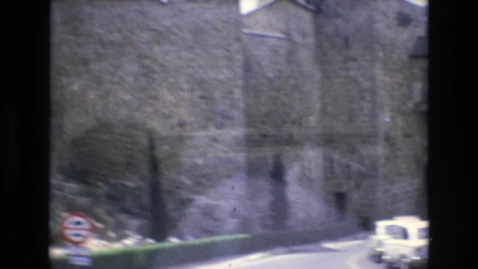 1972 Spain 8mm frame with a large old stone wall, curving roadway, roadside sign, and small light-colored car near the edge of the walled city street.