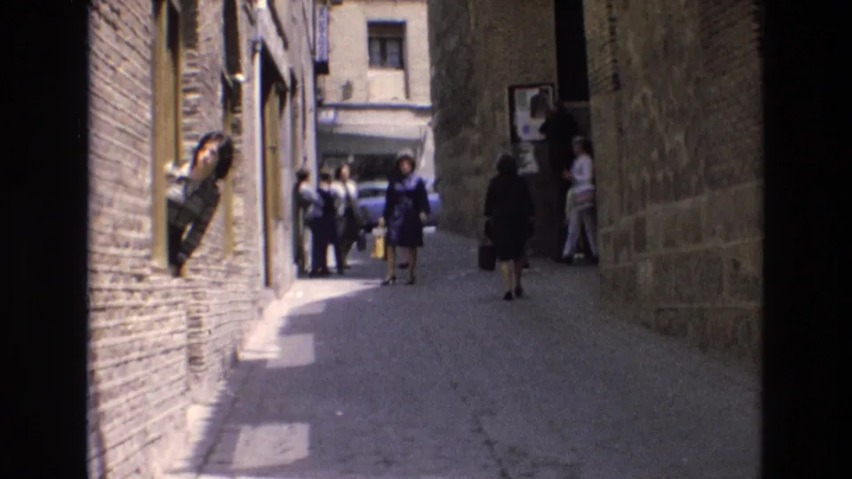 1972 Spain 8mm frame looking down a narrow cobblestone alley between stone and brick walls, with pedestrians gathered ahead and a woman leaning from a window.