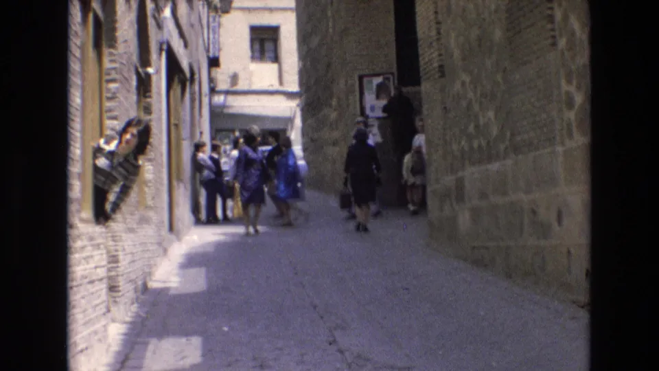 1972 Spain 8mm frame of people walking and gathering in a narrow walled alley, with a woman leaning from a window and sunlit cobblestones in the foreground.