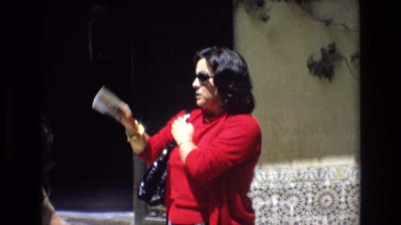 In 1972 Spain home-movie footage, a woman in a red sweater and dark sunglasses holds a small booklet near patterned tilework and a shadowed doorway.