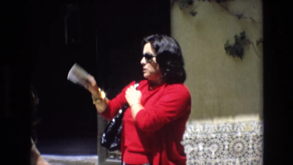 In 1972 Spain home-movie footage, a woman in a red sweater and dark sunglasses holds a small booklet near patterned tilework and a shadowed doorway.