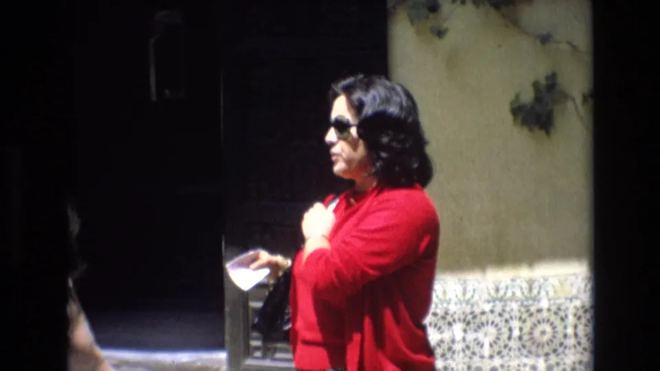 A woman in a red sweater and sunglasses stands near Spanish-style patterned tiles and a dark entrance while handling a small booklet.