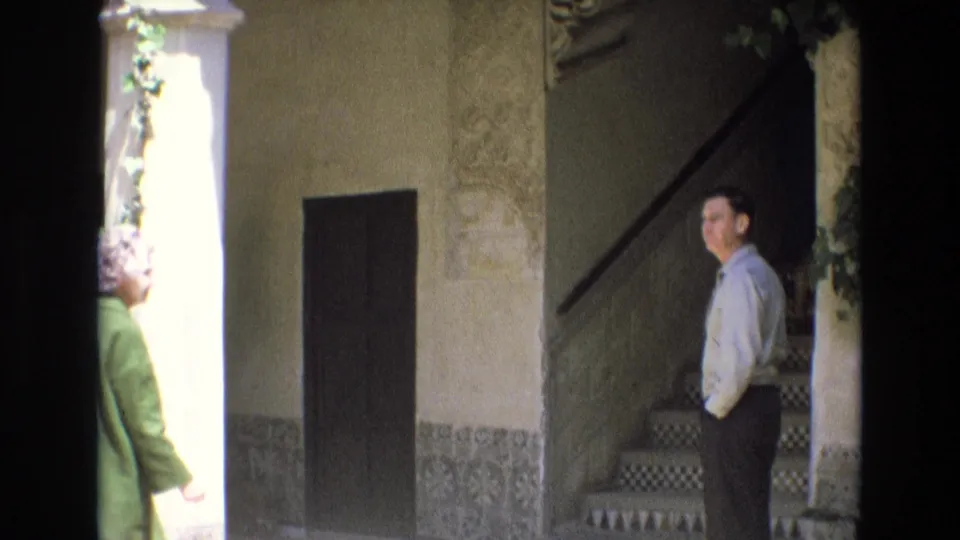 A man in a light shirt stands beside a stairway inside or near a historic Spanish building while a visitor in a green coat appears at the edge of the frame.
