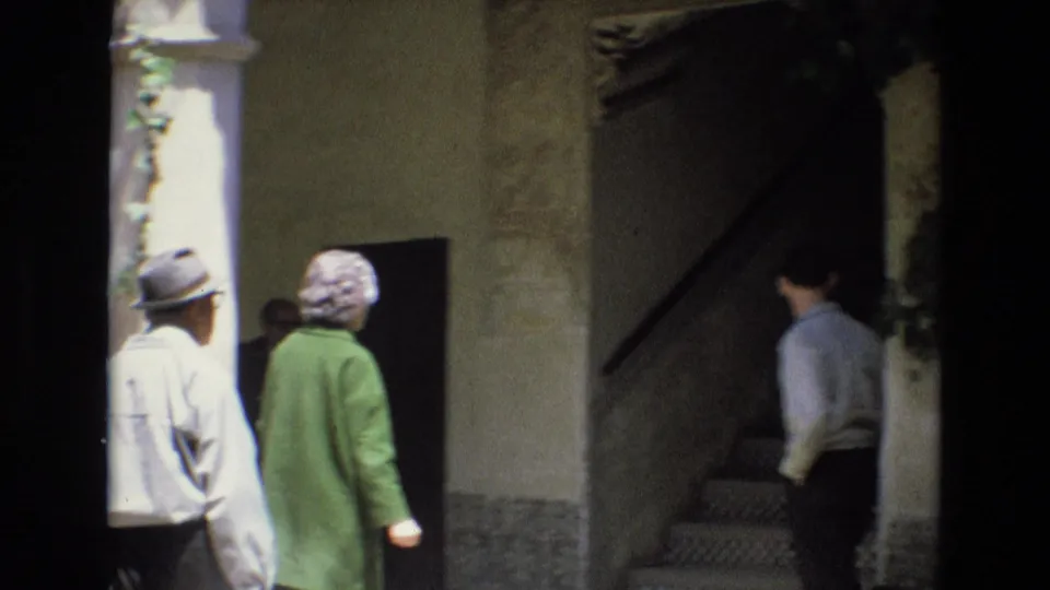 Visitors in coats and hats walk toward a stairway within a Spanish architectural setting, with patterned tiles, stucco walls, and a dark stairwell visible.