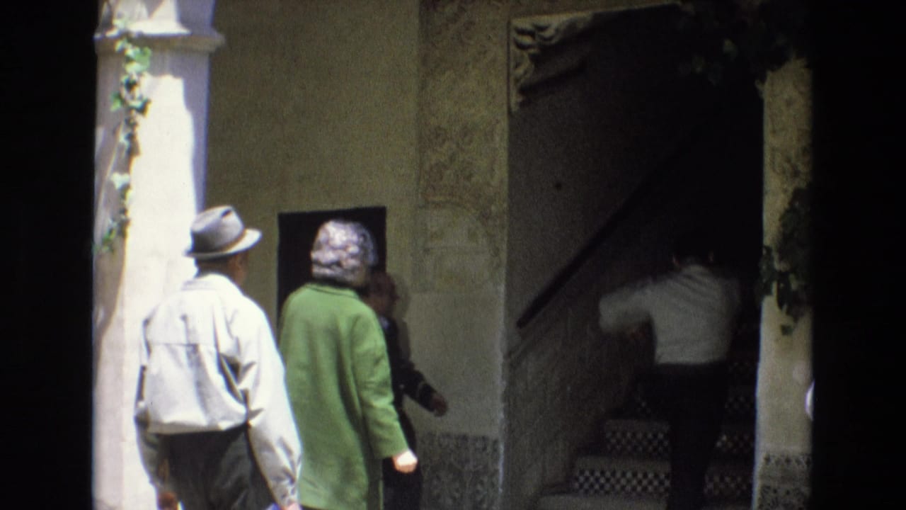In 1972 Spain, a man in a brimmed hat and a woman in a green coat approach a shaded entrance while another person climbs a tiled stair inside the historic building.