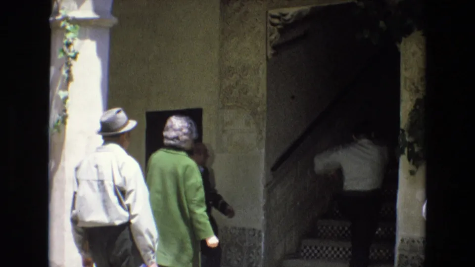 In 1972 Spain, a man in a brimmed hat and a woman in a green coat approach a shaded entrance while another person climbs a tiled stair inside the historic building.