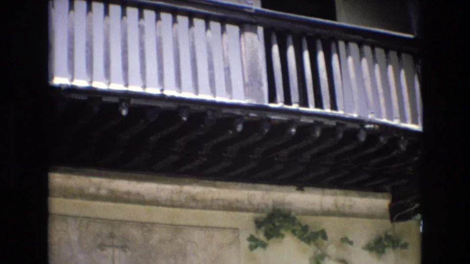 The camera frames a traditional wooden balcony from below, showing its slatted railing, dark support beams, carved wall paneling, and ivy against the building facade.