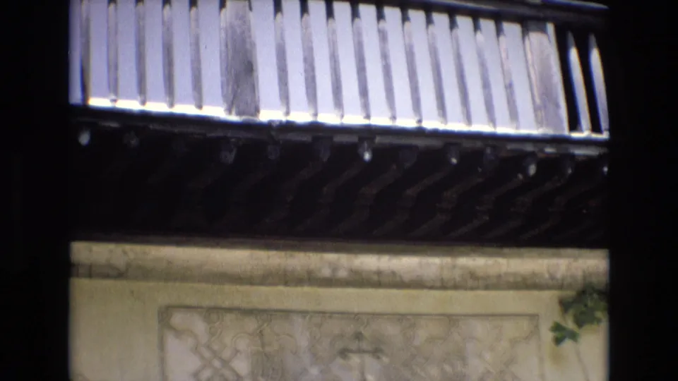 The final frame holds on the balcony and facade, with vertical wooden slats above carved stone decoration, ivy, and a visible cross motif in the wall panel below.