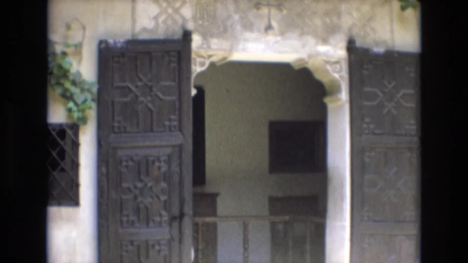 1972 Spain 8mm frame with open carved wooden doors, a stone archway, pale decorative wall surfaces, and a shaded interior passage beyond.