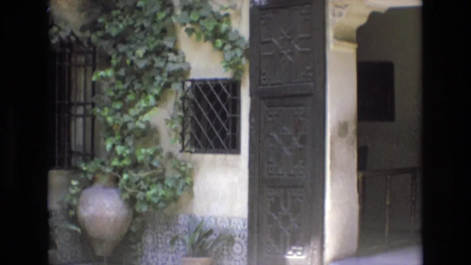 1972 Spain 8mm frame of the courtyard doorway area, with ivy, barred windows, potted plants, carved wooden panels, patterned tiles, and a shaded interior beyond the arch.