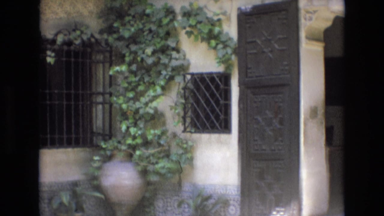 1972 Spain 8mm frame showing a stucco courtyard wall with iron window grilles, ivy, a large terracotta pot, and decorative tilework along the base.